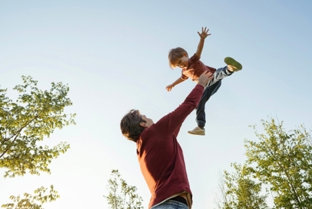 Father and son playing in the park together, dad tossing his happy laughing boy high in the air, having fun and active family time. Happy childhood or Fathers day concept. Space for text