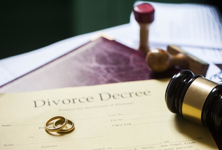 A divorce decree sits on top of a brown leather portfolio. Two golden wedding bands and a wooden gavel sit on top of the divorce decree.
