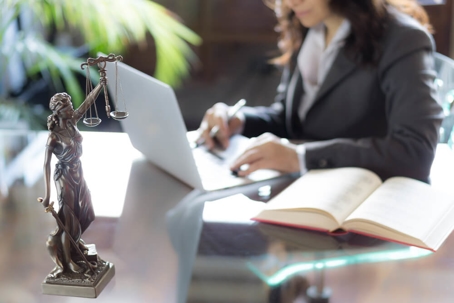 An female attorney sits at a conference table working on a white laptop. A law book and a model of Lady Justice site on the table.
